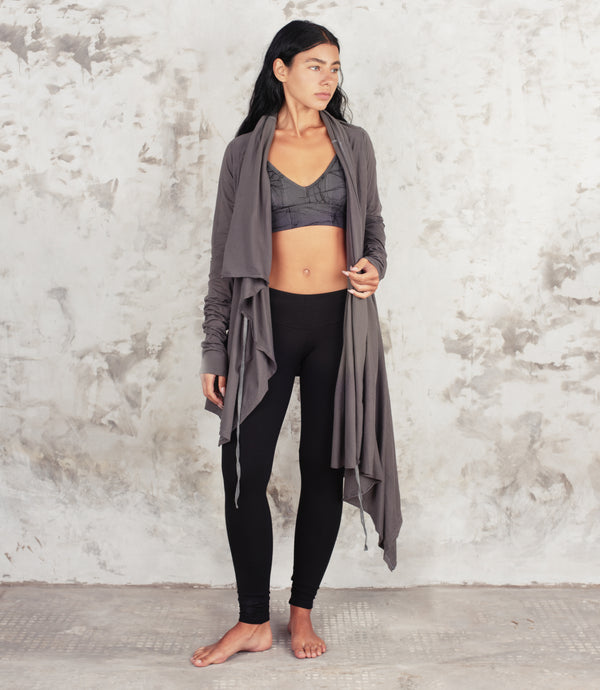 A woman stands barefoot against a textured light gray wall, wearing a long open-front gray cardigan over a gray sports bra and black leggings.