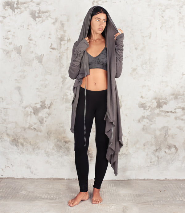 A woman stands barefoot against a textured light gray wall, wearing a long dark gray hooded zip-front cardigan draped over a gray sports bra and black leggings.